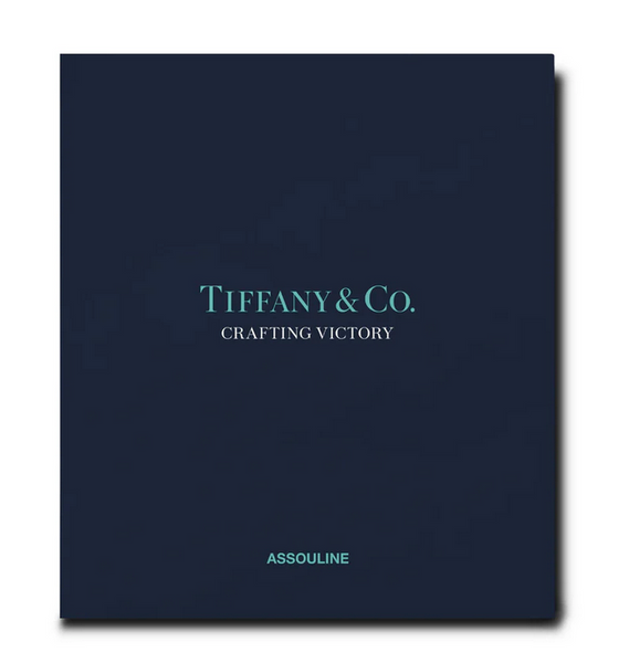 A navy blue book cover titled "Tiffany & Co.: Crafting Victory" in light blue text highlights the artistry of Tiffany hollowware, while the brand name "Assouline" is elegantly printed at the bottom, celebrating silversmithing excellence.