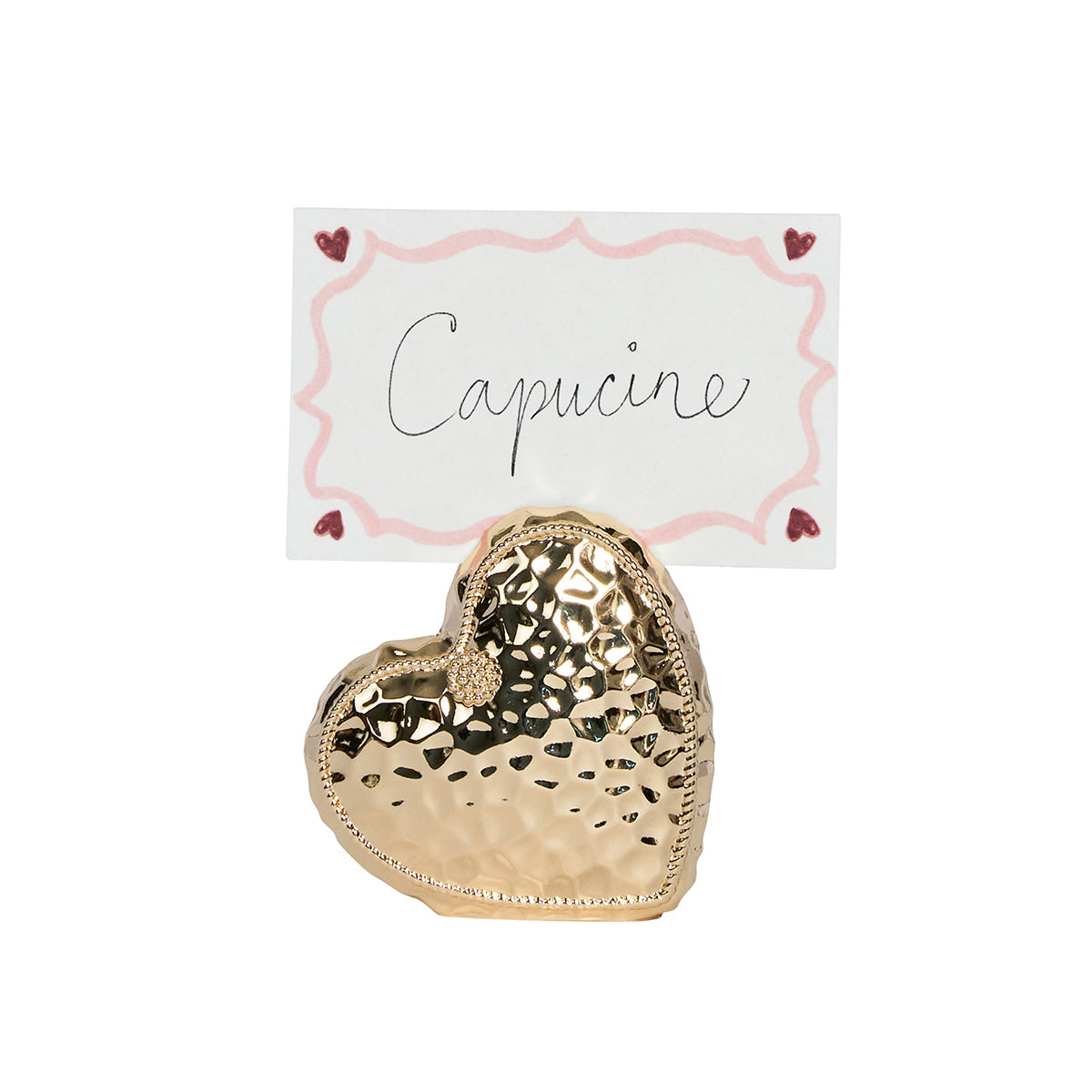 A Berry & Thread Heart Napkin Ring and Place Card Holder by Juliska in metallic gold, with a hammered finish, displays a handwritten "Capucine" card bordered in pink and small hearts.