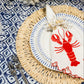 A table setting with a blue and white patterned tablecloth, woven placemat, striped plate, Haute Home’s Red Lobster Napkin Set, cutlery, blue glass, and a floral centerpiece brings seaside charm.