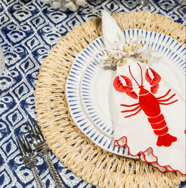 A table setting with a blue and white patterned tablecloth, woven placemat, striped plate, Haute Home’s Red Lobster Napkin Set, cutlery, blue glass, and a floral centerpiece brings seaside charm.