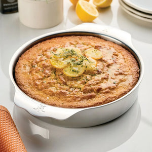A round lemon cake with lemon slices and herbs sits in a Hestan Provisions OvenBond Round Baker on a reflective surface, with plates and halved lemons behind. The baker is oven and broiler safe for versatile baking.