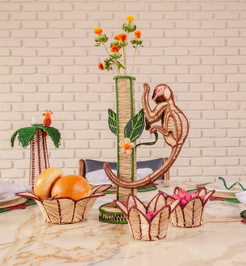 A welcoming table centerpiece showcases the Mercedes Salazar Monkey Bloom Mico Flower Vase filled with flowers, encircled by natural raffia baskets containing bread and pink items, all arranged on a marble table before a white brick wall for an artisanal dining display.