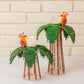 Add a handcrafted touch to your dining decor with the Mercedes Salazar Hoatzin Palm Candle Holder & Flower Vase set—two woven holders adorned with leaf accents and vibrant parrot designs, each holding battery-operated votive candles and showcased against a white brick wall backdrop.