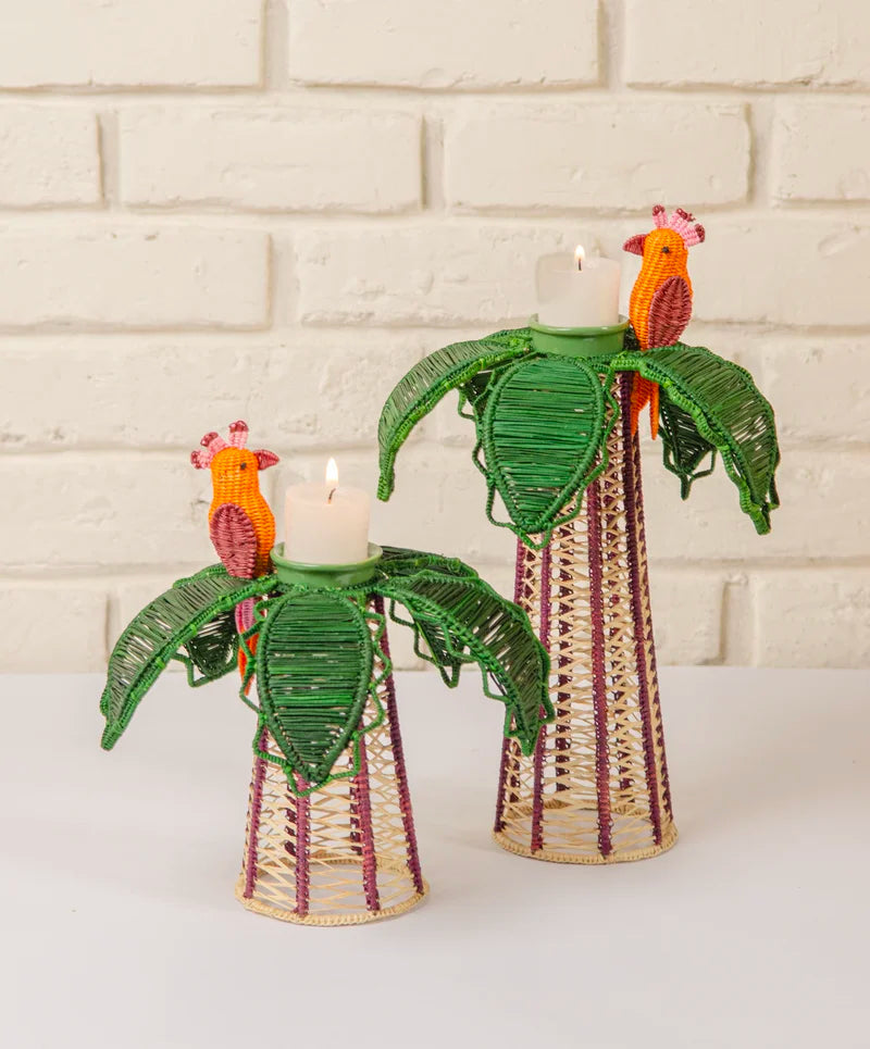 Add a handcrafted touch to your dining decor with the Mercedes Salazar Hoatzin Palm Candle Holder & Flower Vase set—two woven holders adorned with leaf accents and vibrant parrot designs, each holding battery-operated votive candles and showcased against a white brick wall backdrop.