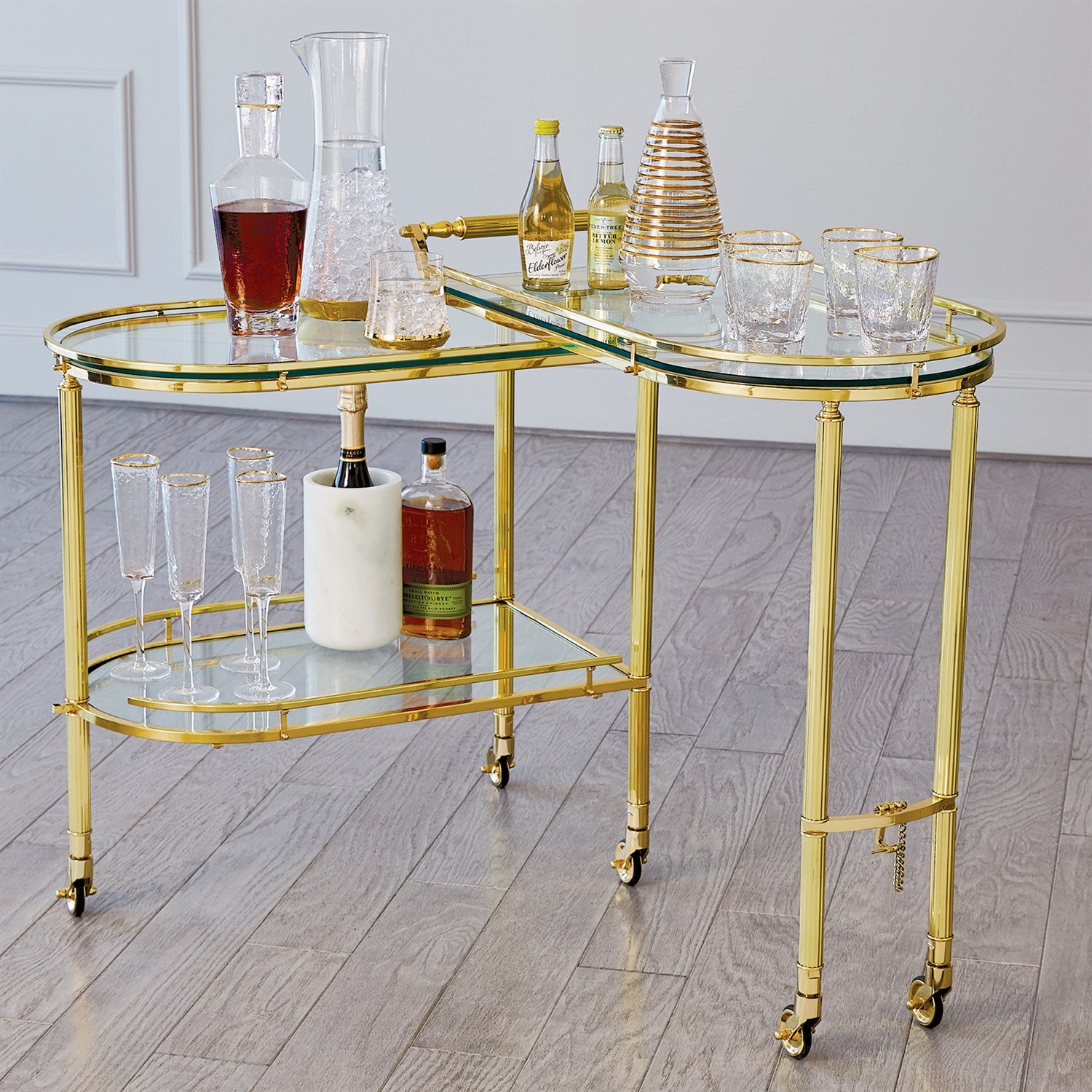 The Gallery Folding Bar Shiny Brass by Global Views showcases decanters, bottles, glasses, and an ice bucket on a light wood floor in a modern room.