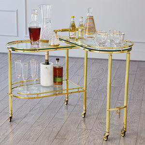 The Gallery Folding Bar Shiny Brass by Global Views showcases decanters, bottles, glasses, and an ice bucket on a light wood floor in a modern room.