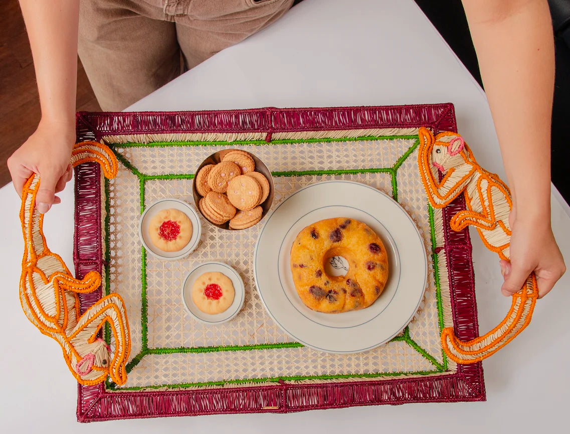 A person holds the Mercedes Salazar Monkey Jungle Party Tray, which is adorned with two bowls of pudding, a plate of cookies, and a plate with a round cake on a white table, creating an inviting dining setting.
