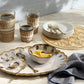 A marble tabletop set with woven glasses, stacked plates and forks, a round Blue Pheasant Cleo Placemat with braided edges, a dish of lemon wedges, oysters on ice, and a cloth napkin.