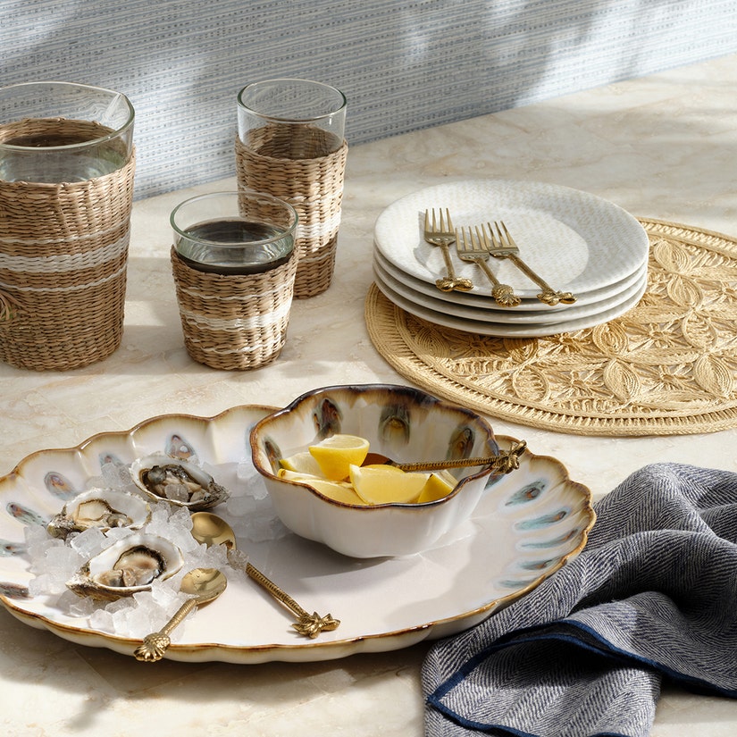 A marble tabletop set with woven glasses, stacked plates and forks, a round Blue Pheasant Cleo Placemat with braided edges, a dish of lemon wedges, oysters on ice, and a cloth napkin.
