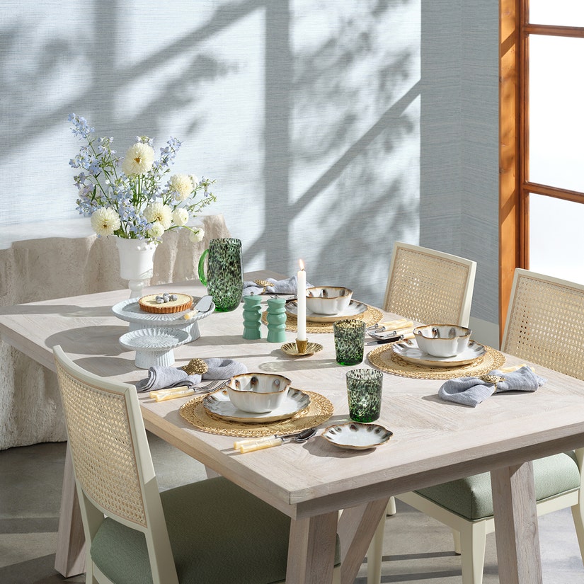 A dining table set for four features Blue Pheasant's Soleil Placemats, complemented by plates, bowls, napkins, green glasses, a candle, a floral centerpiece, textured decor accents, and sunlight streaming through the window.