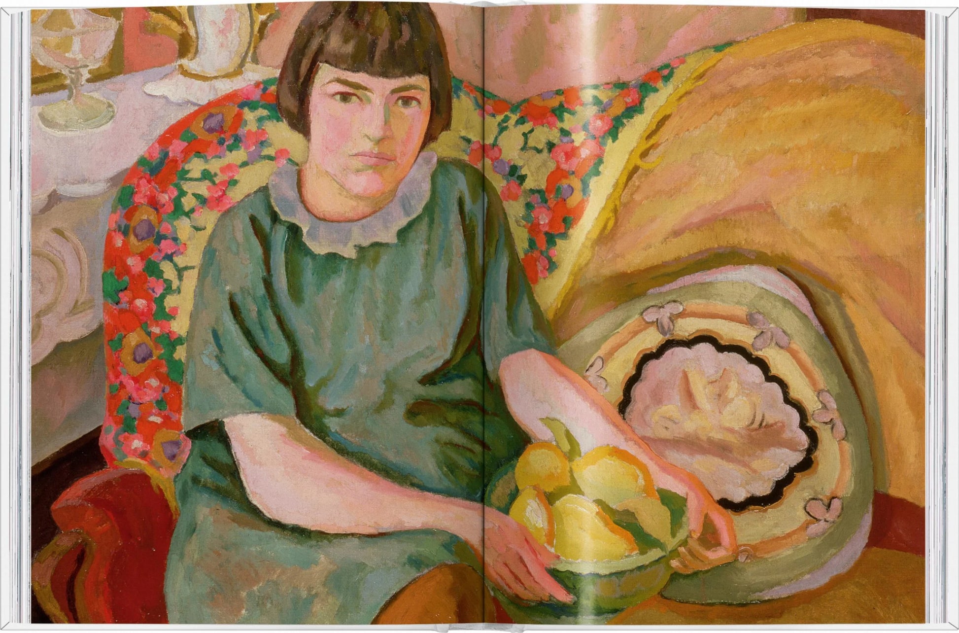 A woman with short dark hair in a green dress sits on a patterned chair, holding Taschen’s “The Gourmand's Lemon: A Collection of Stories and Recipes”—a serene moment where food, literature, and art beautifully intertwine.