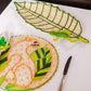 Two woven placemats from Mercedes Salazar—one the Frog Leaf Amazonas Tray, shaped like a leaf with a frog motif, and the other round with an animal outlined in orange—add charm to this dining setting. Crafted from raffia fiber, they rest beside a knife and napkin on a white surface, bringing a natural touch to your table.