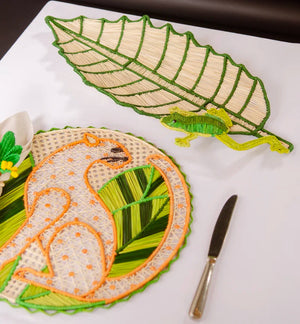 Two woven placemats from Mercedes Salazar—one the Frog Leaf Amazonas Tray, shaped like a leaf with a frog motif, and the other round with an animal outlined in orange—add charm to this dining setting. Crafted from raffia fiber, they rest beside a knife and napkin on a white surface, bringing a natural touch to your table.