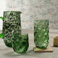 Three green textured glass vessels—a pitcher and two tumblers—are arranged on a flat surface with a Blue Pheasant Jasby Coaster Set, set against pale floral wallpaper.