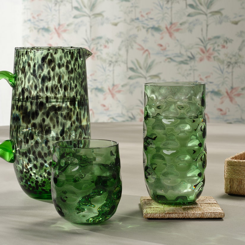 Three green textured glass vessels—a pitcher and two tumblers—are arranged on a flat surface with a Blue Pheasant Jasby Coaster Set, set against pale floral wallpaper.