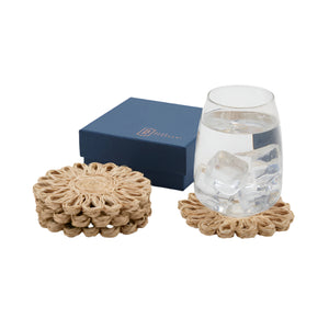 A glass of water with ice rests on a Teigan Natural Floral Coaster by Blue Pheasant, beside a stack of matching handcrafted coasters and the set’s closed blue box.