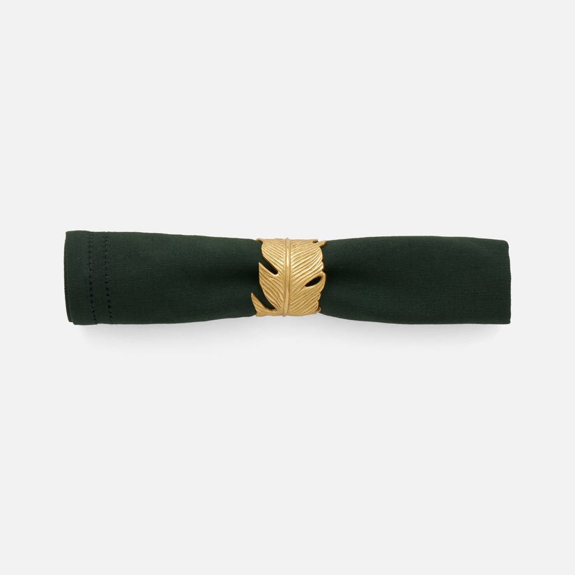 A dark green cloth napkin is rolled and secured with a Blue Pheasant Neela Napkin Ring, featuring a gold brass feather design, displayed on a plain white background.