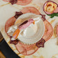 A table setting features a white plate with a pink bird-shaped napkin ring, a flower-shaped bowl on a marble surface, and a handwoven raffia placemat from the Lily & Heliconia Placemat Collection by Mercedes Salazar.