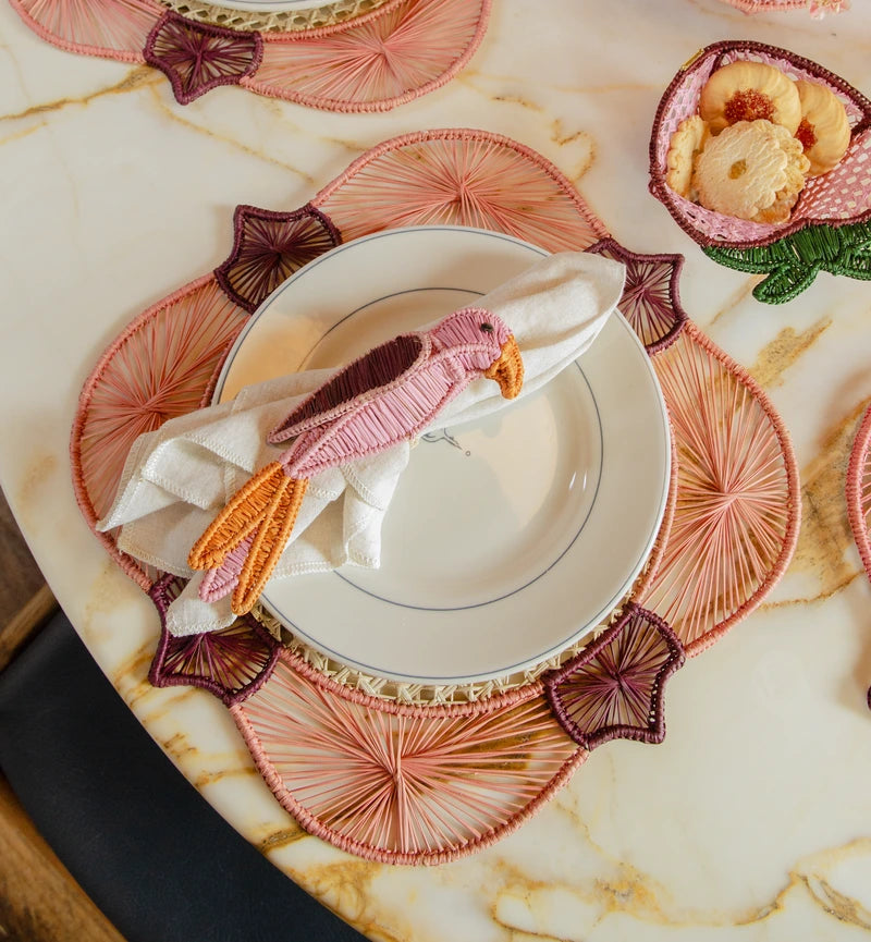 A table setting features a white plate with a pink bird-shaped napkin ring, a flower-shaped bowl on a marble surface, and a handwoven raffia placemat from the Lily & Heliconia Placemat Collection by Mercedes Salazar.