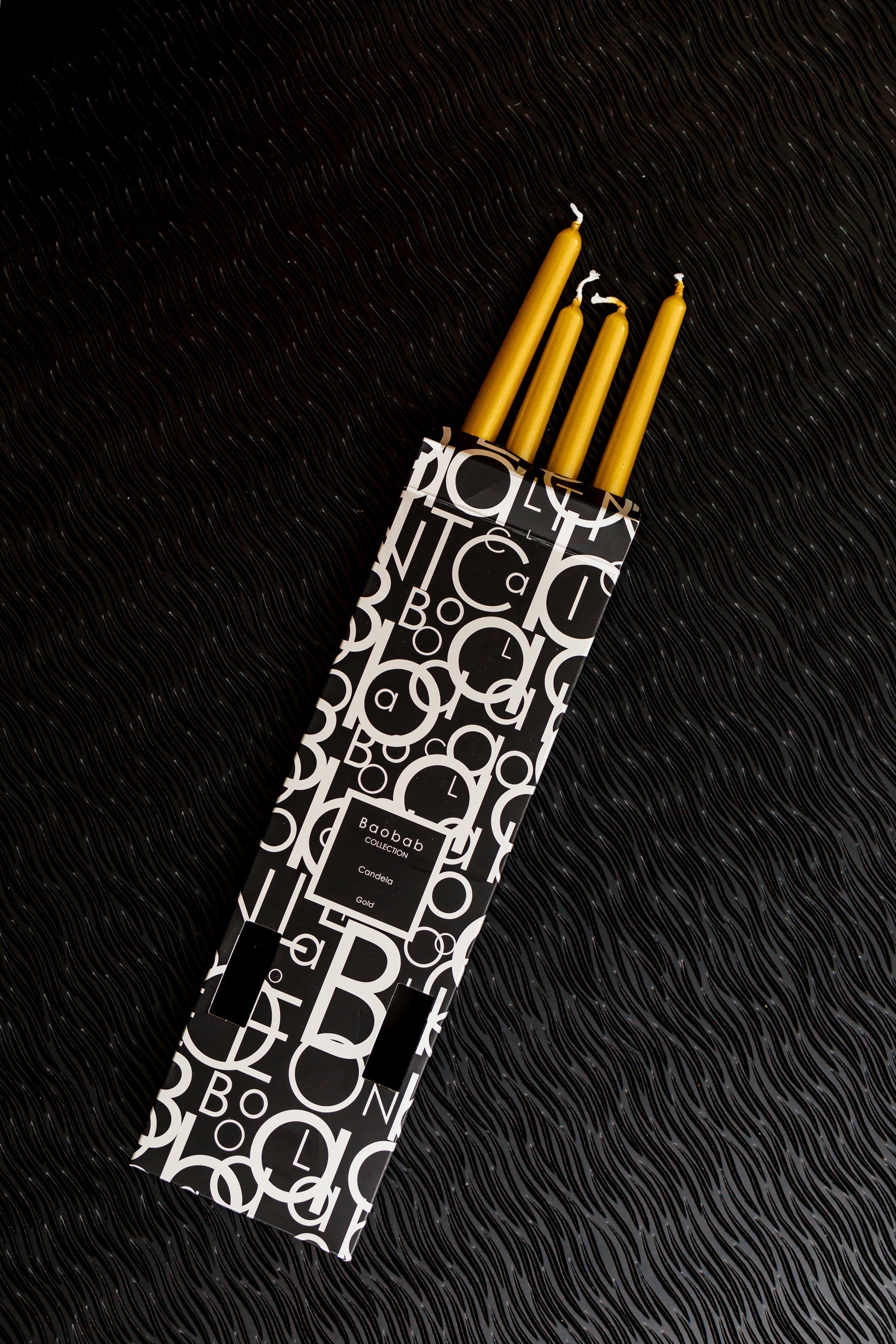 A box of Baobab Collection Candela Candlesticks, featuring sophisticated black and white packaging, rests elegantly on a dark textured surface, making it ideal for enhancing your favorite candlestick. The glaze exudes sophistication while ensuring an extended burning time for any occasion.