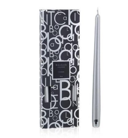A silver tapered candle is displayed next to its box, adorned with a black and white letter pattern design, ready to elegantly complement any candlestick from the Baobab Collection Candela by Baobab.