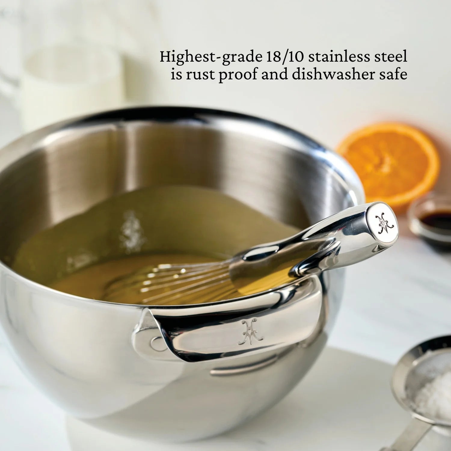 A Hestan Provision Balloon Whisk rests in a stainless steel bowl beside milk, an orange slice, and sugar. Made from highest-grade 18/10 stainless steel—rust proof and dishwasher safe.