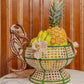 The Forest Muse Mono Bowl by Mercedes Salazar is a woven basket featuring bananas and a pineapple, adorned with floral patterns and finished with a charming monkey-shaped handle. Exquisitely handcrafted from raffia, this playful decor piece—reminiscent of Salazar’s signature style—rests on a marble surface against a wooden wall, bringing the spirit of Bogotá, Colombia into your home.