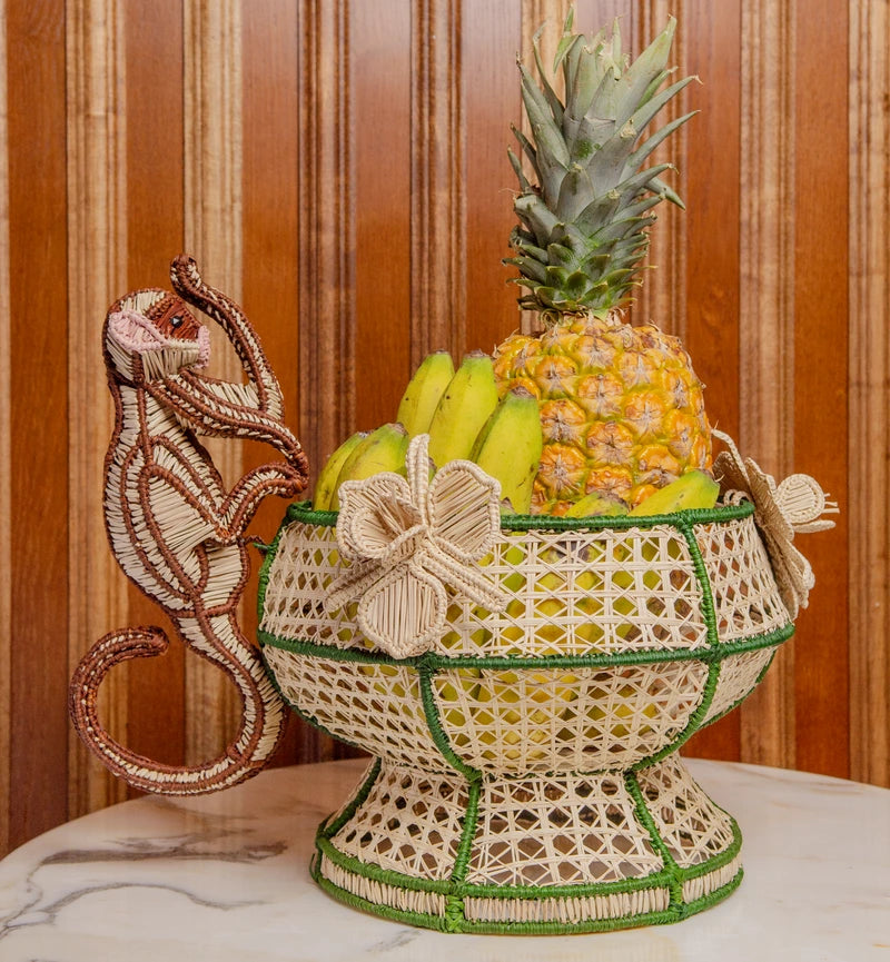 The Forest Muse Mono Bowl by Mercedes Salazar is a woven basket featuring bananas and a pineapple, adorned with floral patterns and finished with a charming monkey-shaped handle. Exquisitely handcrafted from raffia, this playful decor piece—reminiscent of Salazar’s signature style—rests on a marble surface against a wooden wall, bringing the spirit of Bogotá, Colombia into your home.