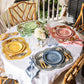 A round table for four is set on the porch with Juliska’s Scallop Stripe Placemat Collection, colorful plates, napkins, silverware, glasses, and a white floral centerpiece for a charming tablescape.