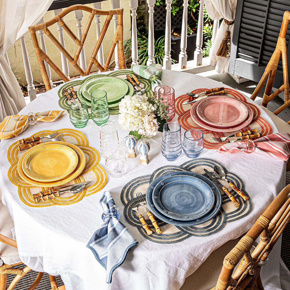A round table for four is set on the porch with Juliska’s Scallop Stripe Placemat Collection, colorful plates, napkins, silverware, glasses, and a white floral centerpiece for a charming tablescape.