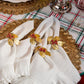 Four white Mademoiselle Napkins by Juliska with ruffle borders and gold and red berry napkin rings are arranged on a gold placemat over a plaid tablecloth to create a romantic tablescape.