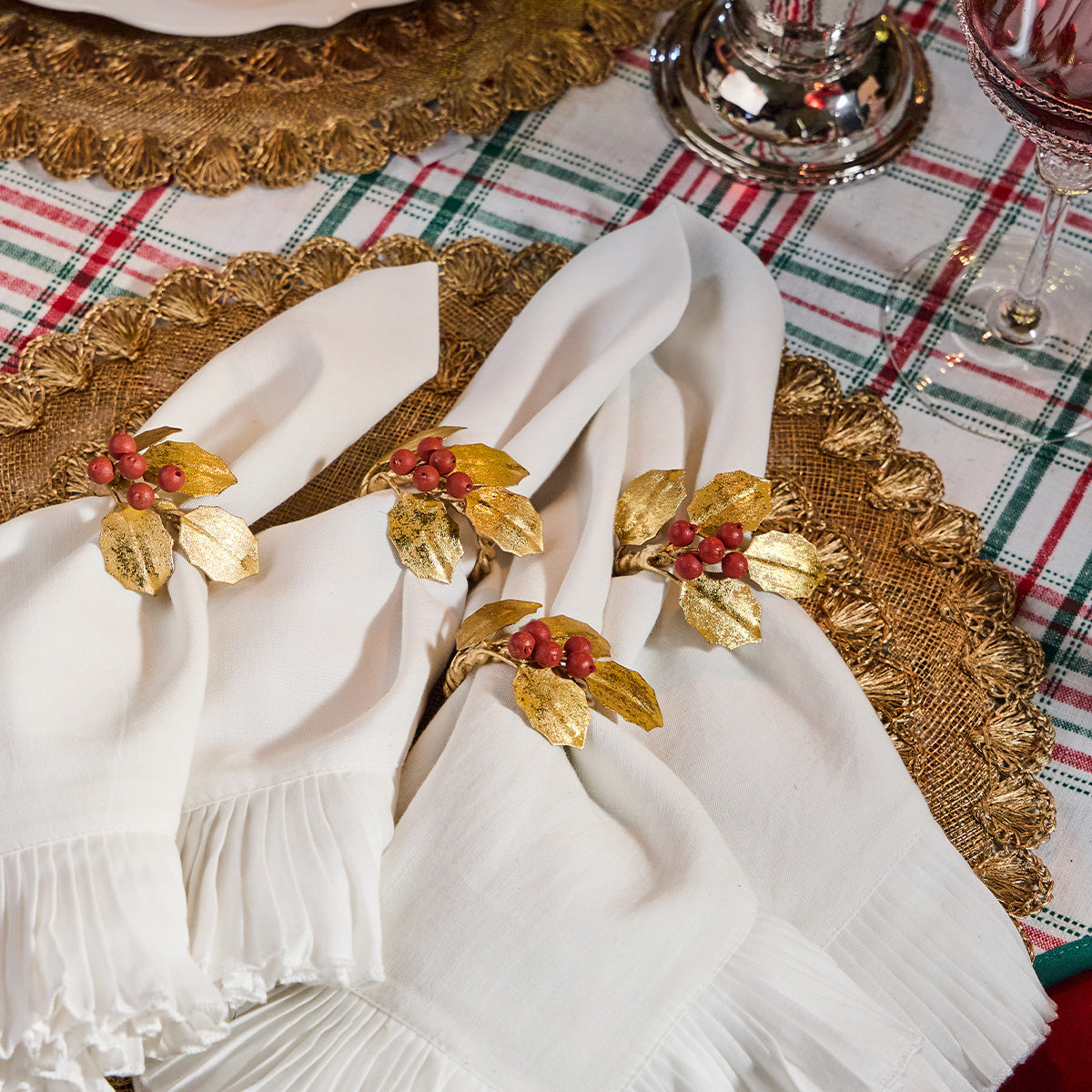 Four white Mademoiselle Napkins by Juliska with ruffle borders and gold and red berry napkin rings are arranged on a gold placemat over a plaid tablecloth to create a romantic tablescape.
