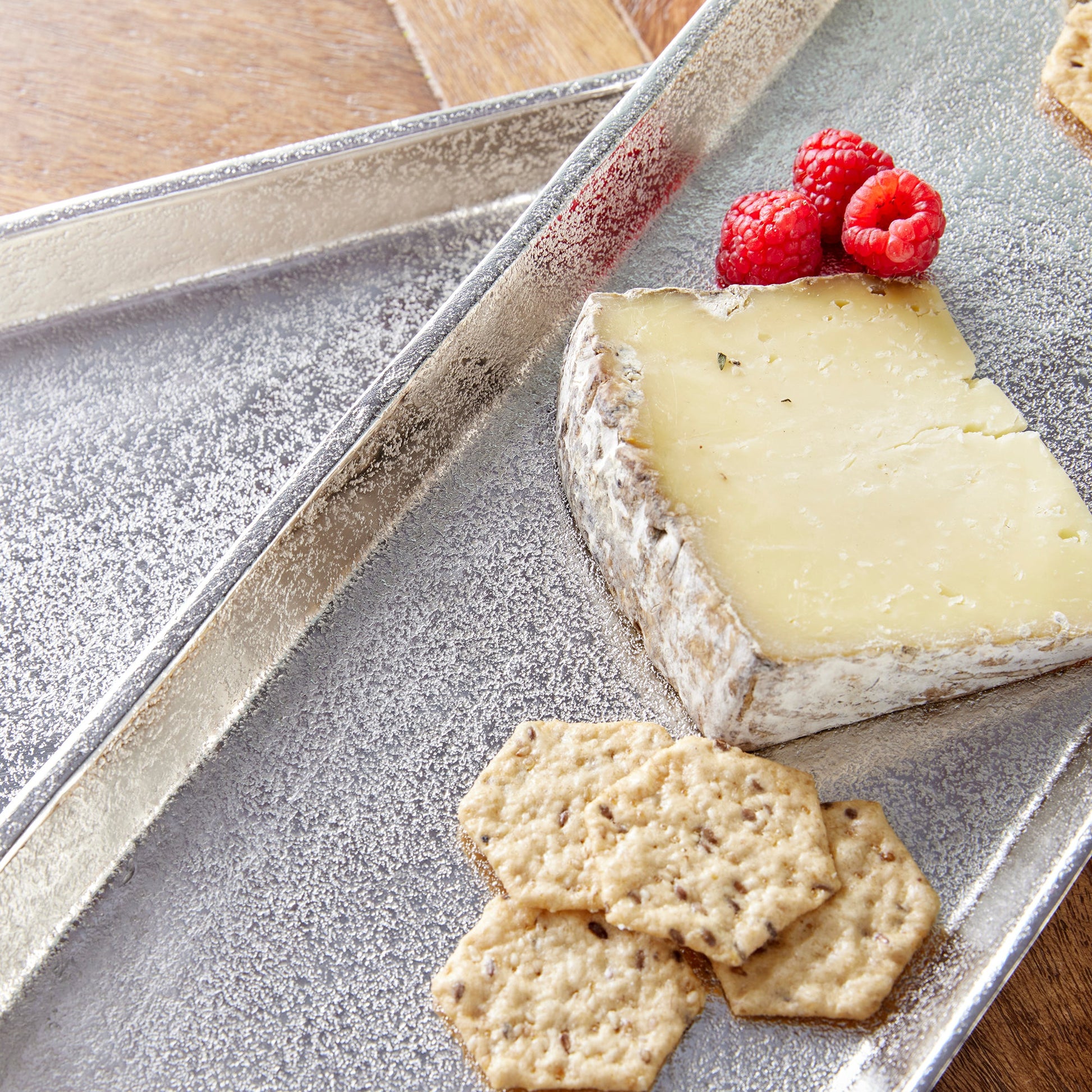 A wedge of cheese with rind, three hexagonal crackers, and three raspberries are artfully arranged on the Helena Long Tray Collection by Napa Home & Garden for a designer look.