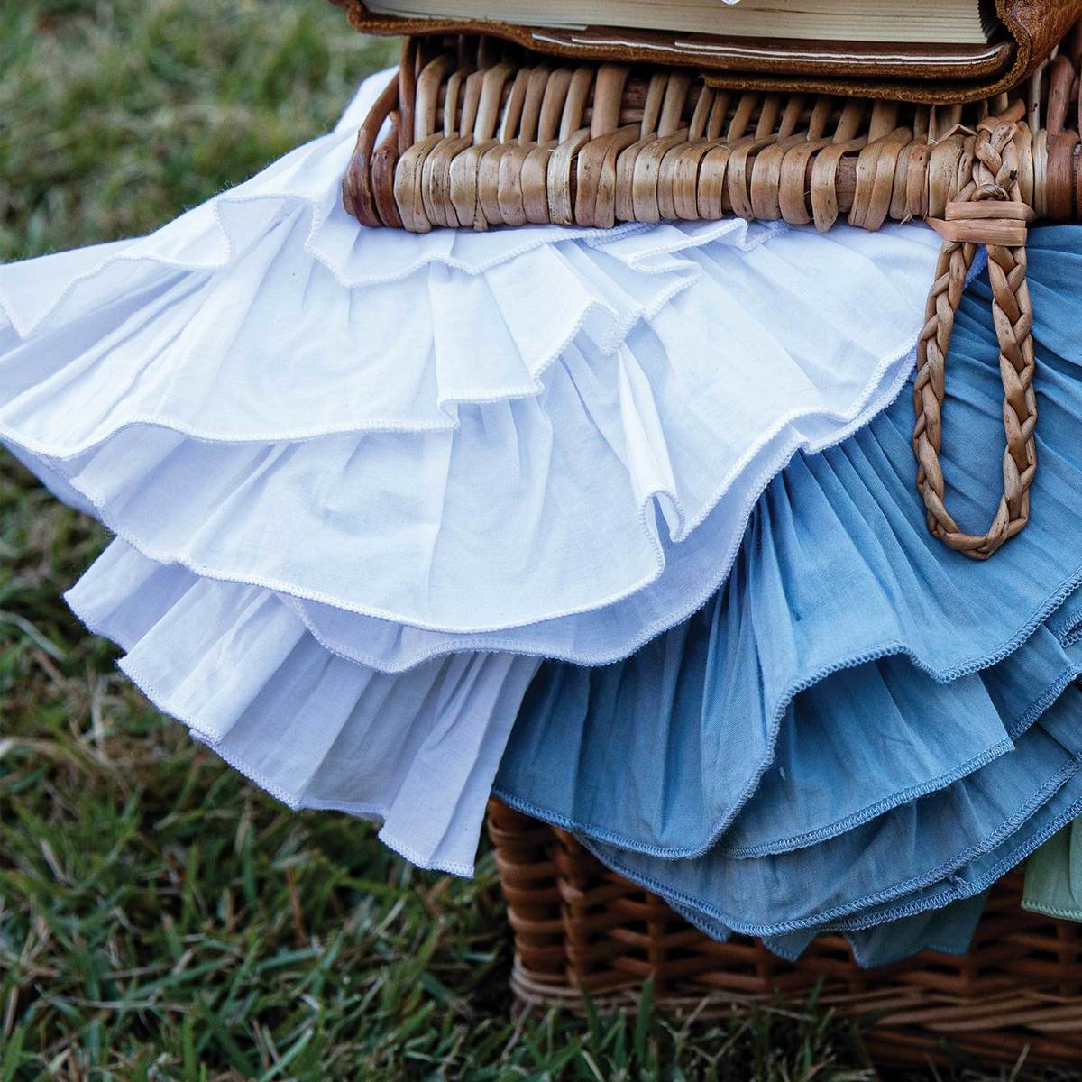 Juliska’s Mademoiselle Napkin Set features white and blue napkins with knife-pleat ruffle borders, elegantly draped over a wicker basket with grass in the background.