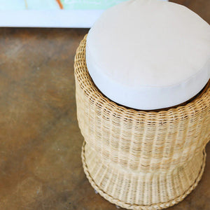 The Varnish Collection Belle Stool features a cylindrical wicker base and a round cream upholstered seat, offering cozy seating. Photographed from above, it's shown on a concrete floor.