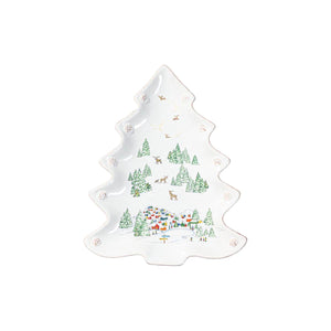 The Juliska Berry & Thread North Pole Tree Platter Collection features a white ceramic platter shaped like a Christmas tree, adorned with a winter village scene of trees, houses, and reindeer in green, red, and blue.