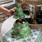 A hand lifts the lid of the Juliska Berry & Thread Basil Christmas Tree Cookie Jar, filled with treats and set on a festive tablecloth beside green mugs.