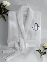 A folded white Milagro Robe by Matouk, set of 2 for Keaton and Alexis's Registry, with a belt and "JSM" initials in navy blue, surrounded by white flowers and green leaves.