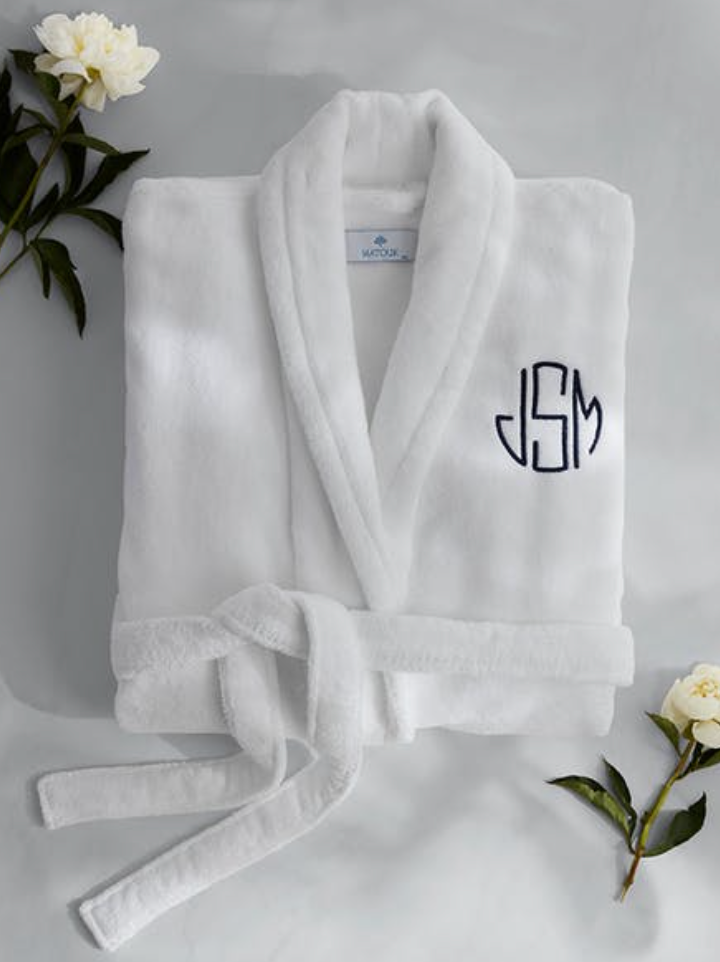 A folded white Milagro Robe by Matouk, set of 2 for Keaton and Alexis's Registry, with a belt and "JSM" initials in navy blue, surrounded by white flowers and green leaves.
