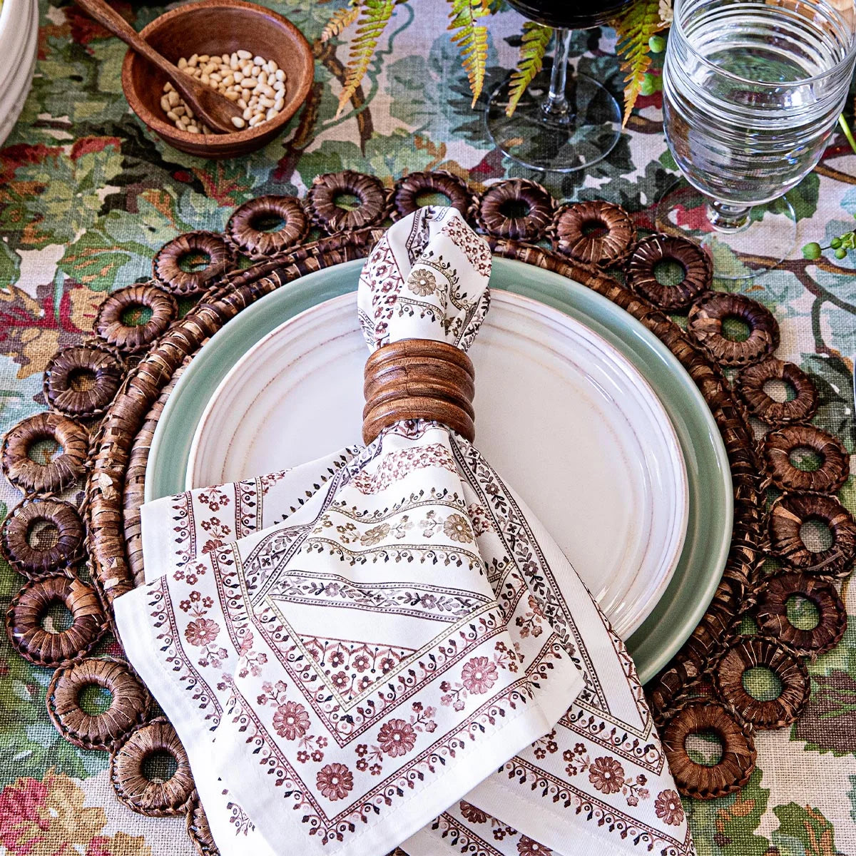 A table setting inspired by the Spanish countryside, featuring a Juliska Seville Stripe Napkin in a wooden ring atop a plate, surrounded by a woven placemat and complemented by a glass of water.
