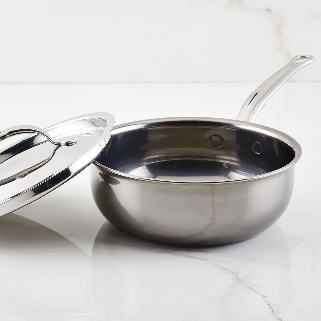 A Hestan NanoBond Titanium Stainless Steel Saucier Pan, 2qt, with its lid beside it rests on a white marble surface; it's designed to be safe for use with metal utensils.