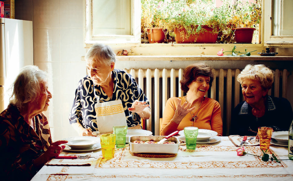 Four elderly women sit around a table in a sunlit kitchen adorned with potted plants on the windowsill, sharing laughter and heartfelt stories as they prepare delicious meals inspired by Pasta Grannies: Comfort Cooking from Chronicle Books.