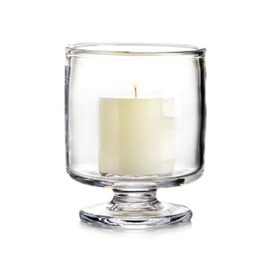 A white pillar candle sits inside a clear, stemmed Simon Pearce Nantucket Hurricane Collection holder against a white background.