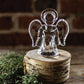 The Simon Pearce Angel Collection features a clear handmade angel figurine displayed on a round wooden platform with green foliage nearby, set against a brick wall background.