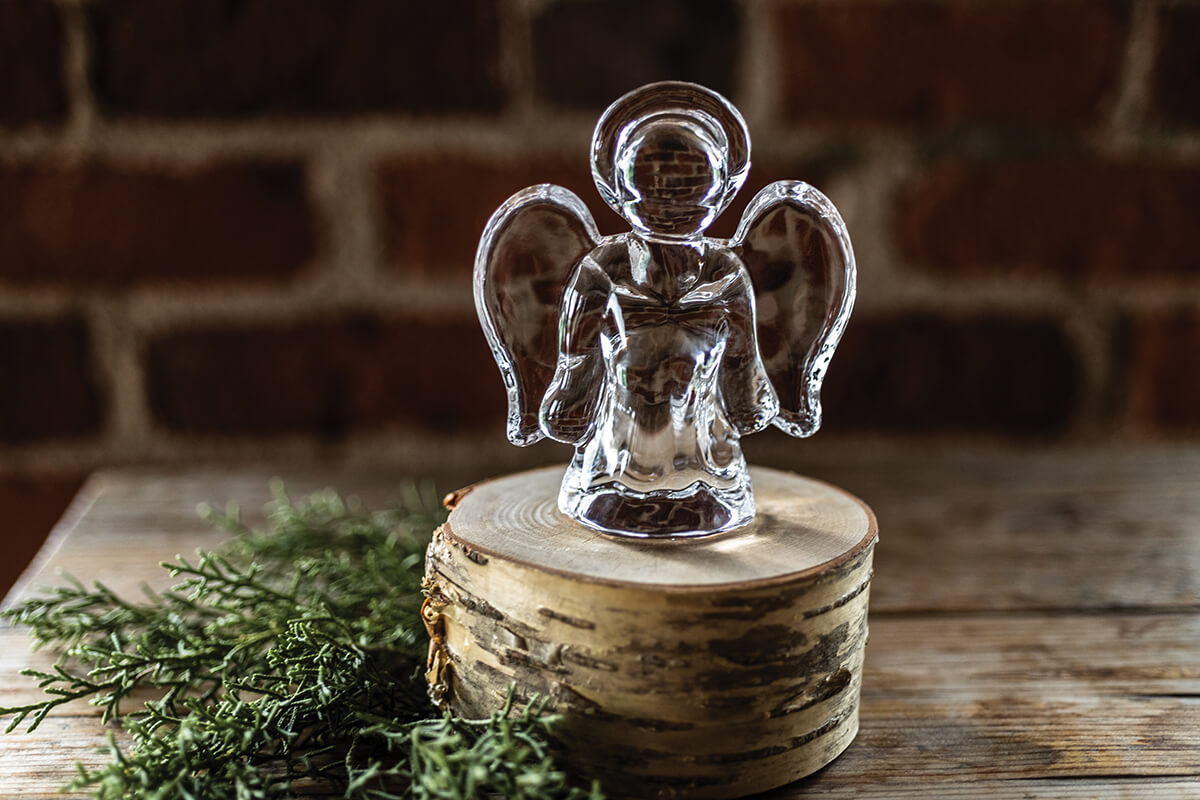 The Simon Pearce Angel Collection features a clear handmade angel figurine displayed on a round wooden platform with green foliage nearby, set against a brick wall background.
