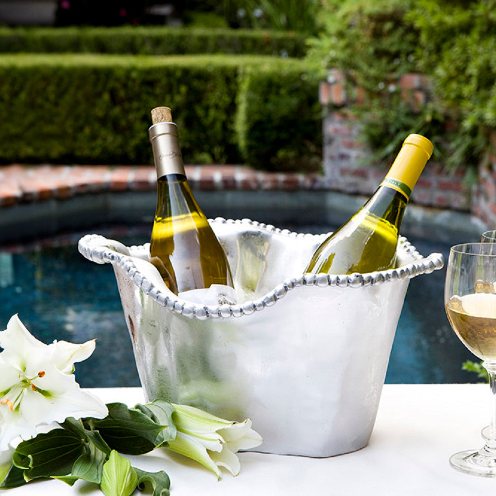 Two bottles of white wine chill in a Beatriz Ball Organic Pearl Ice Bucket Collection, set on a table with flowers and wine glasses, overlooking a serene pool and lush garden.