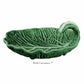 The Bordallo Pinheiro Cabbage Leaf Green Collection features a green ceramic dish resembling a curled leaf with a wavy edge. Handmade with care, it includes the text: "With Curvature 7".