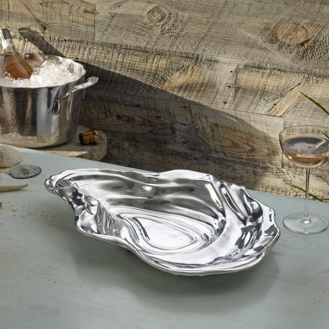 The Beatriz Ball Ocean Oyster Bowl Collection, featuring a hand-polished silver oyster shell-shaped serving platter, sits on a light blue table beside a wine glass, seashells, and an ice bucket with rosé.