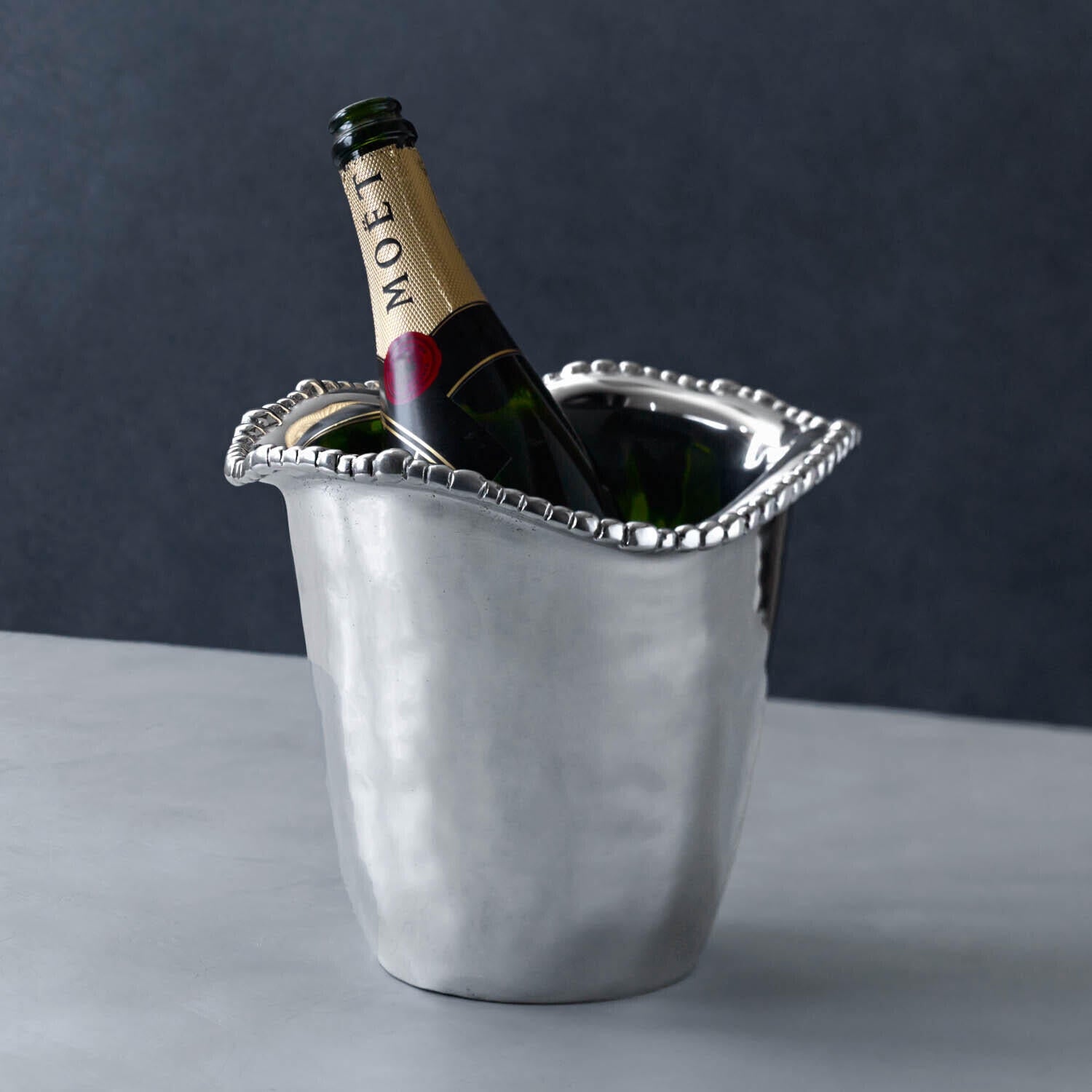 A bottle of Moët champagne rests in a Beatriz Ball Organic Pearl Ice Bucket Collection piece, featuring a wavy beaded rim, on a gray surface against a dark background.