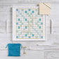 The Scrabble Bianco Edition by WS Game Company features a wood-framed board with tiles spelling "PLAY," "BIG," "OCEAN," "BREEZE," "SHORE," and "ELEGANT." A blue tile bag, score pads, and sand timer sit beside it on a wooden surface, perfect for an ocean breeze design.